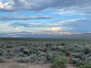 Sagebrush landscape in front of mountains and clouds and multi-colored skycap